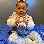 Malaya is registered to the contest to win money with this photo: baby, child, smiling, pink_bow, sitting, shoe, footwear, gray_sweatpants, white_socks, hoodie, indoor, blue_blanket, person, cute, happy, portrait, playful, young_child, cozy, casual_clothing