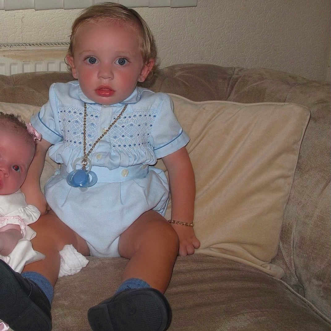 Wallace-Raymond joined the competition — help win amazing prizes! baby, black_shoes, blue_eyes, blue_outfit, bracelet, child, couch, curtain, doll, face, furniture, indoor, pacifier, person, pillow, radiator, sitting, toddler, toy, wall