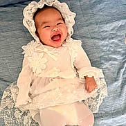 Bela is registered to the contest to win money with this photo: baby, infant, child, smiling, laughing, portrait, bonnet, lace, dress, formalwear, floral, sleeves, tights, lying_down, quilt, blue_background, indoor, happy, face, photography