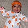 baby, blanket, child, closeup, cute, expression, face, hands, happy, indoors, infant, joyful, lying_down, onesie, orange_hat, portrait, pumpkin_pattern, skin, smiling, warm_clothing