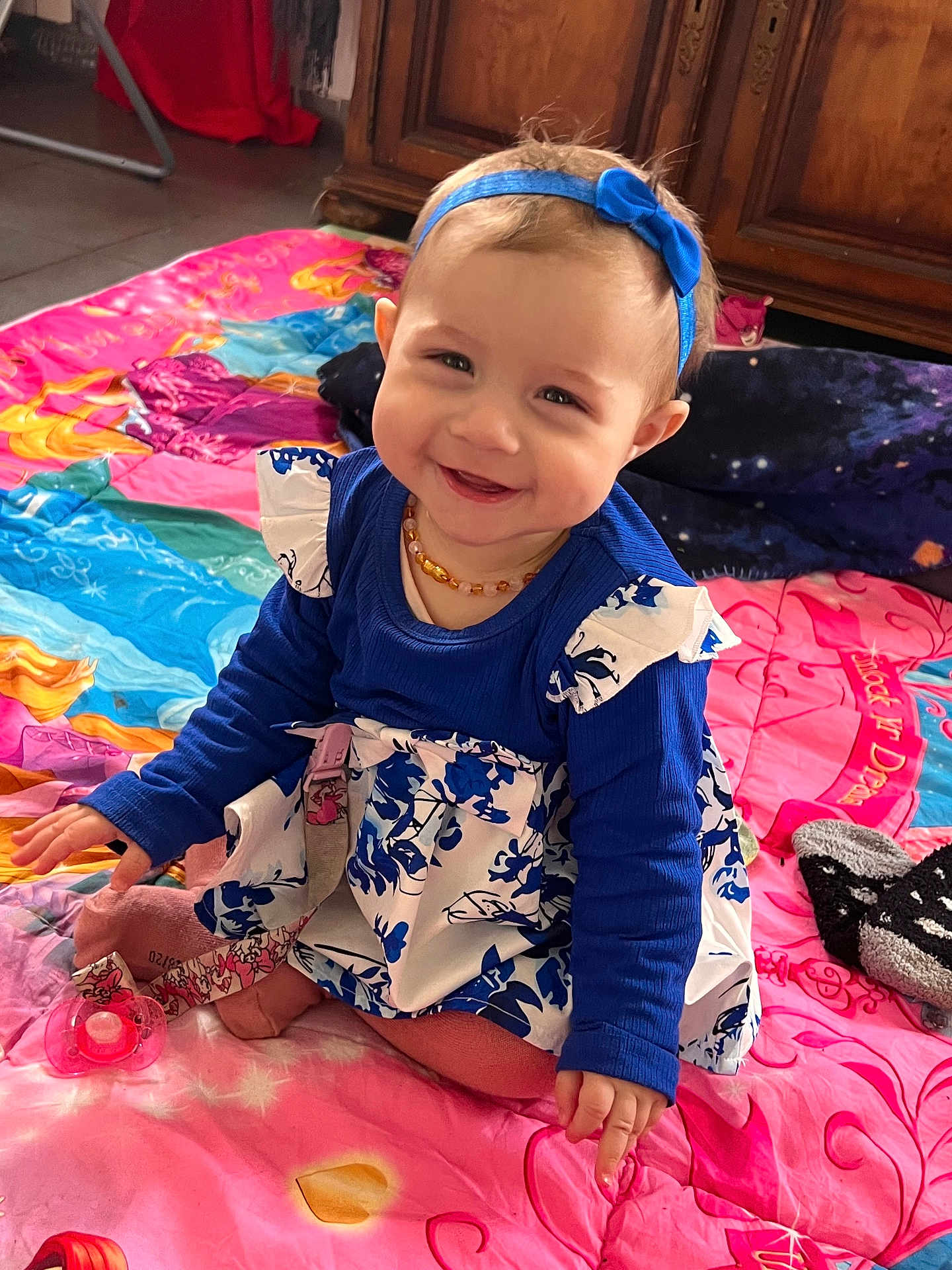 Leana participe au concours pour gagner de l'argent avec cette photo : baby, child, smiling, sitting, blue_headband, dress, floral_pattern, blanket, colorful, pacifier, socks, wooden_furniture, indoor, cute, happy, portrait, floor, playful, headband, clothing