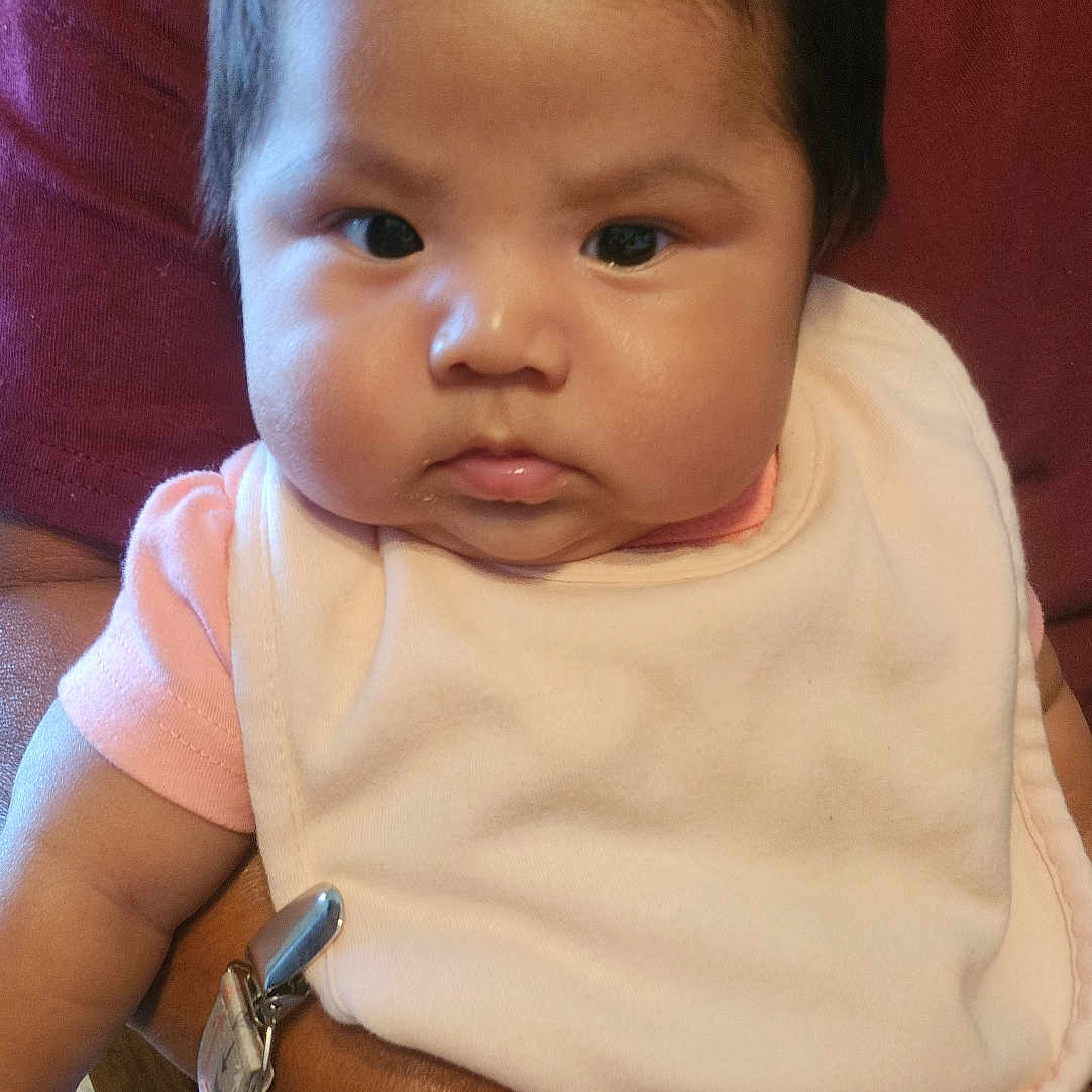 Kaylsha joined the competition — help win amazing prizes! arm, baby, bib, child, chubby_cheeks, clothing, fabric, face, hand, holding, indoor, infant, person, pink_clothing, portrait, seated, serious_expression, short_hair, skin, wooden_chair