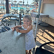 Léa participe au concours pour gagner de l'argent avec cette photo : child, girl, white_dress, smiling, outdoor, table, chair, pool, palm_tree, umbrella, sunlight, summer, sandals, restaurant, vacation, holiday, happy, person, wooden_floor, daytime