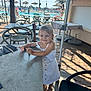 child, girl, white_dress, smiling, outdoor, table, chair, pool, palm_tree, umbrella, sunlight, summer, sandals, restaurant, vacation, holiday, happy, person, wooden_floor, daytime