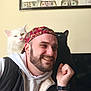 man, smile, cat, white_cat, black_cat, heterochromia, cap, beard, hoodie, vest, indoor, portrait, feline, pet, happy, face, watch, wall_art, money, friendly