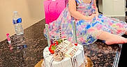 Sally is registered to the contest to win money with this photo: child, girl, birthday, birthday_cake, cake, candles, dress, colorful, portrait, sitting, kitchen, countertop, cabinets, curly_hair, brown_hair, face, celebration, party, water_bottle, indoor