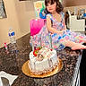 child, girl, birthday, birthday_cake, cake, candles, dress, colorful, portrait, sitting, kitchen, countertop, cabinets, curly_hair, brown_hair, face, celebration, party, water_bottle, indoor
