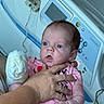 baby, infant, hand, hospital_bed, medical_equipment, pink_clothing, person, newborn, adult_hand, bed_control_panel, curious_expression, indoors, healthcare, closeup, skin, face, holding, hospital, patient, care