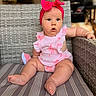 baby, child, pink_bow, headband, pink_outfit, striped_cushion, wicker_chair, indoors, curious_expression, sitting, plush_toy, armrest, chubby_legs, closeup, portrait, casual, cute, infant, furniture, home