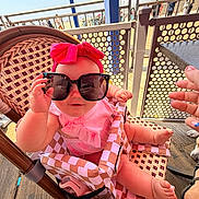 Kamryn is registered to the contest to win money with this photo: baby, child, sunglasses, headband, pink_bow, high_chair, woven_chair, outdoor, sunny, wooden_floor, metal_fence, hand, nail_polish, playful, cute, infant, summer, boardwalk, people_in_background, footwear