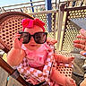 baby, child, sunglasses, headband, pink_bow, high_chair, woven_chair, outdoor, sunny, wooden_floor, metal_fence, hand, nail_polish, playful, cute, infant, summer, boardwalk, people_in_background, footwear