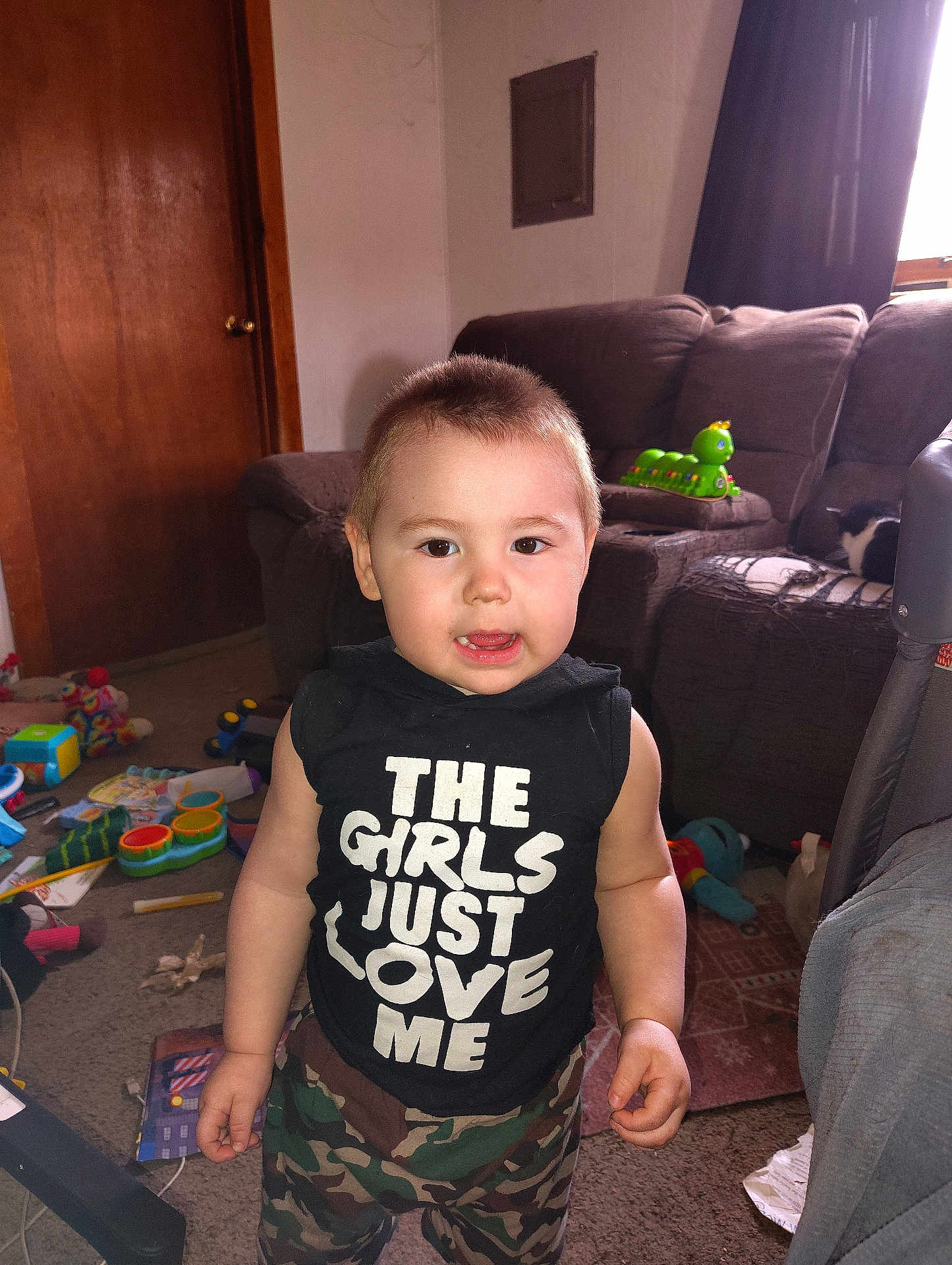 Loki joined the competition — help win amazing prizes! toddler, child, indoor, living_room, camouflage_pants, black_shirt, text_on_clothing, toy, carpet, couch, curtains, door, window, face, person, expression, furniture, messy, cute, home