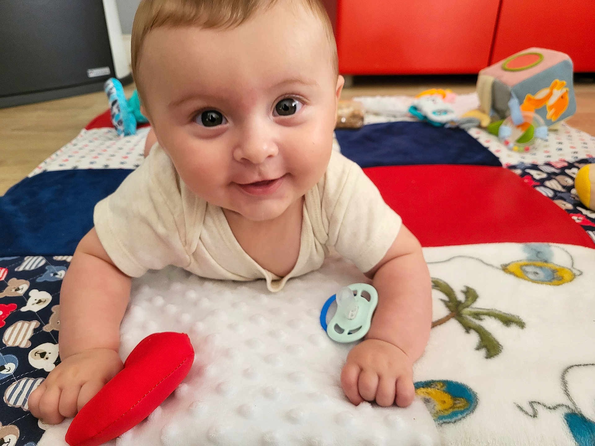 Sacha a rejoint le concours — aidez-le/la à gagner de superbes lots ! baby, smiling, face, infant, toy, pacifier, play_mat, indoor, floor, clothing, hand, colorful, soft_toy, wood_floor, furniture, child, cute, happy, portrait, toddler