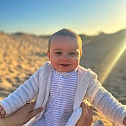 Georgie joined the competition — help win amazing prizes! baby, smiling, child, cozy_clothing, jacket, onesie, hands, beach, sand, sunlight, blue_sky, outdoor, portrait, happy, infant, cute, warm_light, human, person, daylight