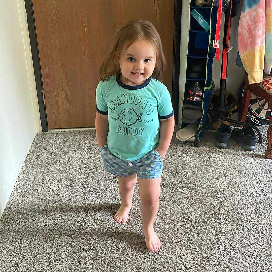 Aurora is registered to the contest to win money with this photo: barefoot, carpet, casual, child, clothing, door, feet, hand_in_pocket, happy, home, indoor, person, shirt, shoe_organizer, shorts, smiling, standing, toddler, wood, young_child