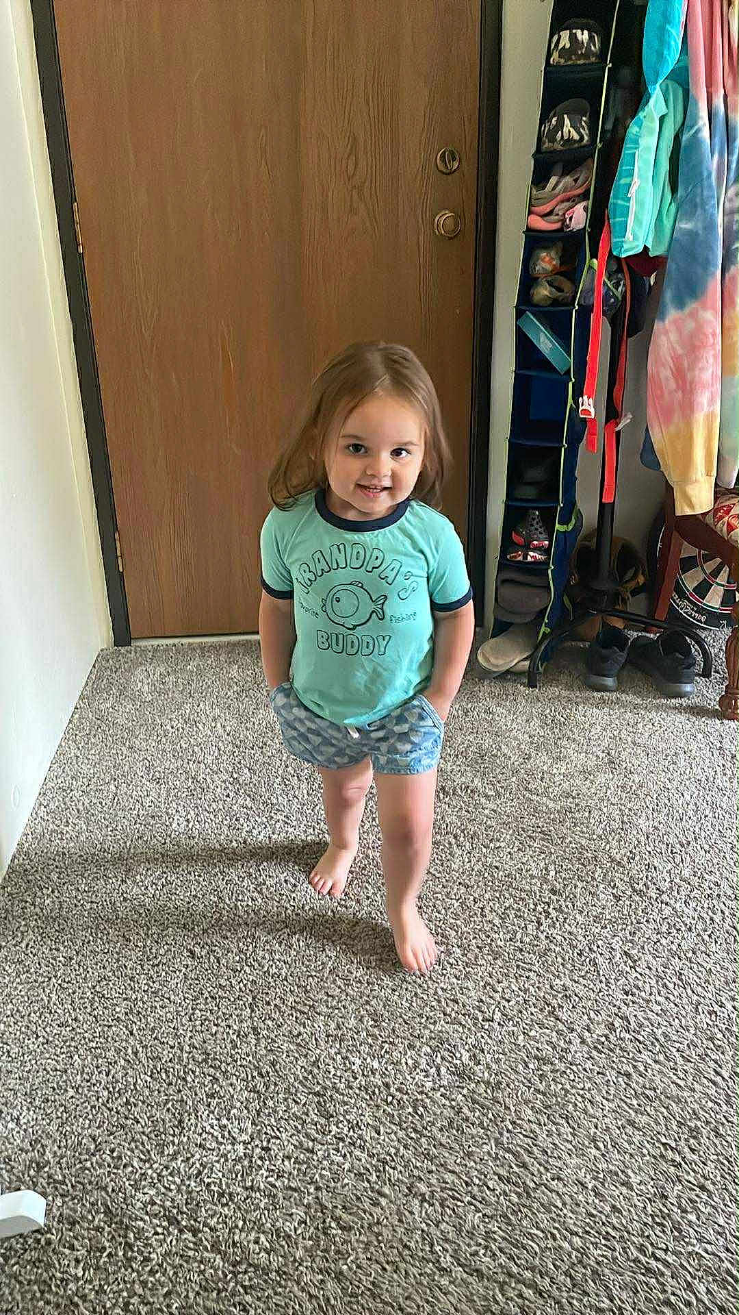 Aurora is registered to the contest to win money with this photo: toddler, child, barefoot, carpet, door, shorts, shirt, smiling, standing, indoor, clothing, shoe_organizer, wood, hand_in_pocket, person, young_child, home, casual, feet, happy
