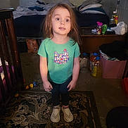 Aurora joined the competition — help win amazing prizes! child, toddler, green_shirt, cat_graphic, standing, rug, indoor, dim_lighting, long_hair, curious_expression, footwear, carpet, bedroom, pillow, table, plastic_bottle, toy, floor, furniture, person