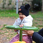 Rita a rejoint le concours — aidez-le/la à gagner de superbes lots ! toddler, child, playground, toy, green_handlebar, yellow_toy, autumn_leaves, outdoor, smiling, white_jacket, black_hat, pom_poms, hand_support, pink_pants, daisy_duck, happy, person, park, fall, cute