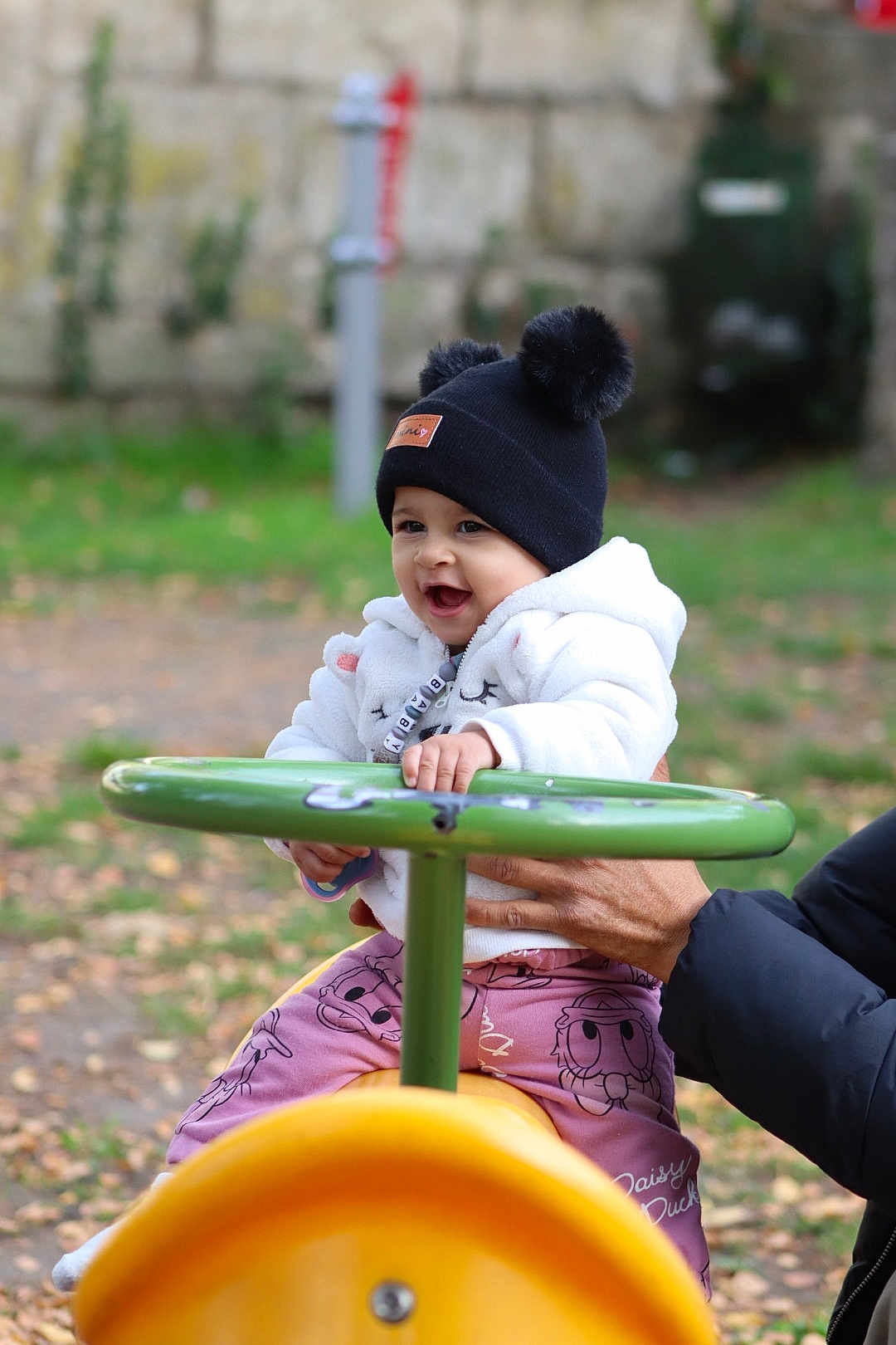 Rita a rejoint le concours — aidez-le/la à gagner de superbes lots ! toddler, child, playground, toy, green_handlebar, yellow_toy, autumn_leaves, outdoor, smiling, white_jacket, black_hat, pom_poms, hand_support, pink_pants, daisy_duck, happy, person, park, fall, cute