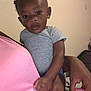 Ace is registered to the contest to win money with this photo: baby, child, indoor, person, holding, pink_shirt, gray_onesie, curly_hair, small_buns, hand, face, looking_at_camera, skin, adult_hand, seated, expression, furniture, wall, casual_clothing, closeup