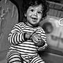 toddler, child, curly_hair, striped_clothing, onesie, smiling, happy, indoor, playpen, baby_toy, black_and_white, portrait, seated, infant, cute, small_hands, face, eyes, expression, young_child