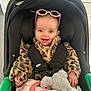 baby, child, car_seat, fuzzy_jacket, leopard_print, pink_sunglasses, stuffed_animal, elephant_toy, blanket, indoor, smiling, happy, person, cute, cozy, infant, seatbelt, clothing, portrait, young_child
