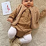baby, infant, milestone_card, clothing, knitted_booties, carpet, soft_texture, brown, beige, cute, smiling, lying_down, portrait, child, indoor, warm_colors, buttoned_shirt, sleeves, white_tights, cozy