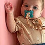baby, pacifier, infant, pink_background, floral_onesie, blue_pants, hand, face, eyes, skin, clothing, cute, lying_down, child, portrait, indoor, soft_lighting, expression, closeup, adorable