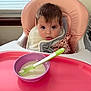 baby, high_chair, pink, purple_bowl, green_spoon, food, feeding, indoor, window, curious, face, child, tongue, bib, clothing, seat_belt, table, expression, meal, infant