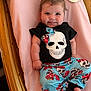 baby, infant, smiling, skull_print, clothing, blue_pants, floral_pattern, bow, pink_blanket, wooden_cradle, cute, happy, child, portrait, indoors, person, laying_down, small_feet, chubby_arms, cozy