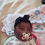 Cimani is registered to the contest to win money with this photo: baby, pacifier, blanket, headband, remote_control, infant, floral_pattern, sleepwear, lying_down, indoor, child, cute, comfort, soft_texture, face, person, young_child, resting, pink, white