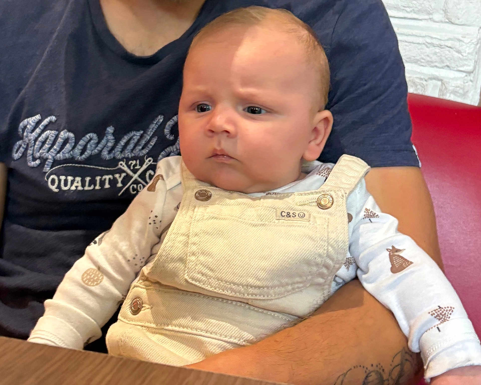 Kaïdo participe au concours pour gagner de l'argent avec cette photo : baby, child, person, infant, overalls, clothing, arm, tattoo, table, sitting, indoor, adult, holding, long_sleeve, pattern, expression, face, skin, casual, seated
