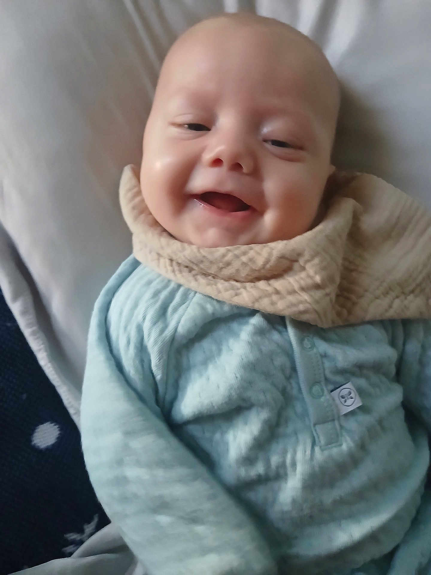 Zander is registered to the contest to win money with this photo: baby, smiling, infant, blue_clothing, bib, bed, blanket, cozy, face, head, closeup, happy, child, person, indoors, soft_texture, cute, young, portrait, resting