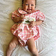 Mia is registered to the contest to win money with this photo: baby, infant, smile, pink_dress, checkered_pattern, lying_down, white_sheet, happy, cute, child, skin, face, hands, legs, toes, short_hair, indoors, portrait, one_person, young_child