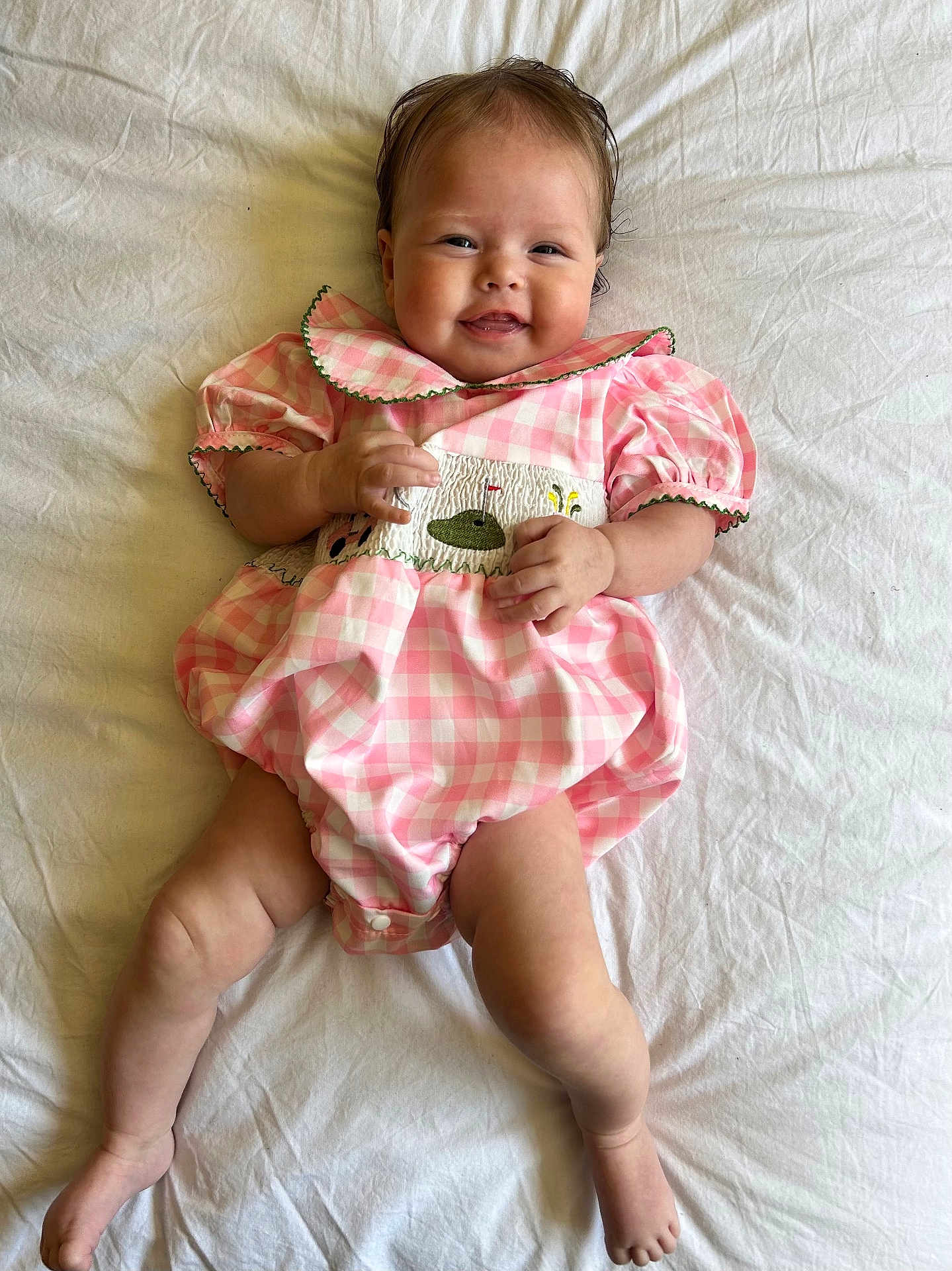 Mia is registered to the contest to win money with this photo: baby, infant, smile, pink_dress, checkered_pattern, lying_down, white_sheet, happy, cute, child, skin, face, hands, legs, toes, short_hair, indoors, portrait, one_person, young_child