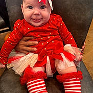 Isabella is registered to the contest to win money with this photo: baby, child, smiling, red_clothing, headband, bow, striped_socks, tulle_skirt, hand, chair, indoor, person, happy, festive, cute, toddler, face, sitting, closeup, holiday