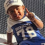 blue_clothing, bowl, boy, cap, casual, child, daylight, finger_gesture, glass_table, happy, outdoor, portrait, relaxed, skin, smiling, sportswear, sunlight, toddler, white_cap, woven_chair