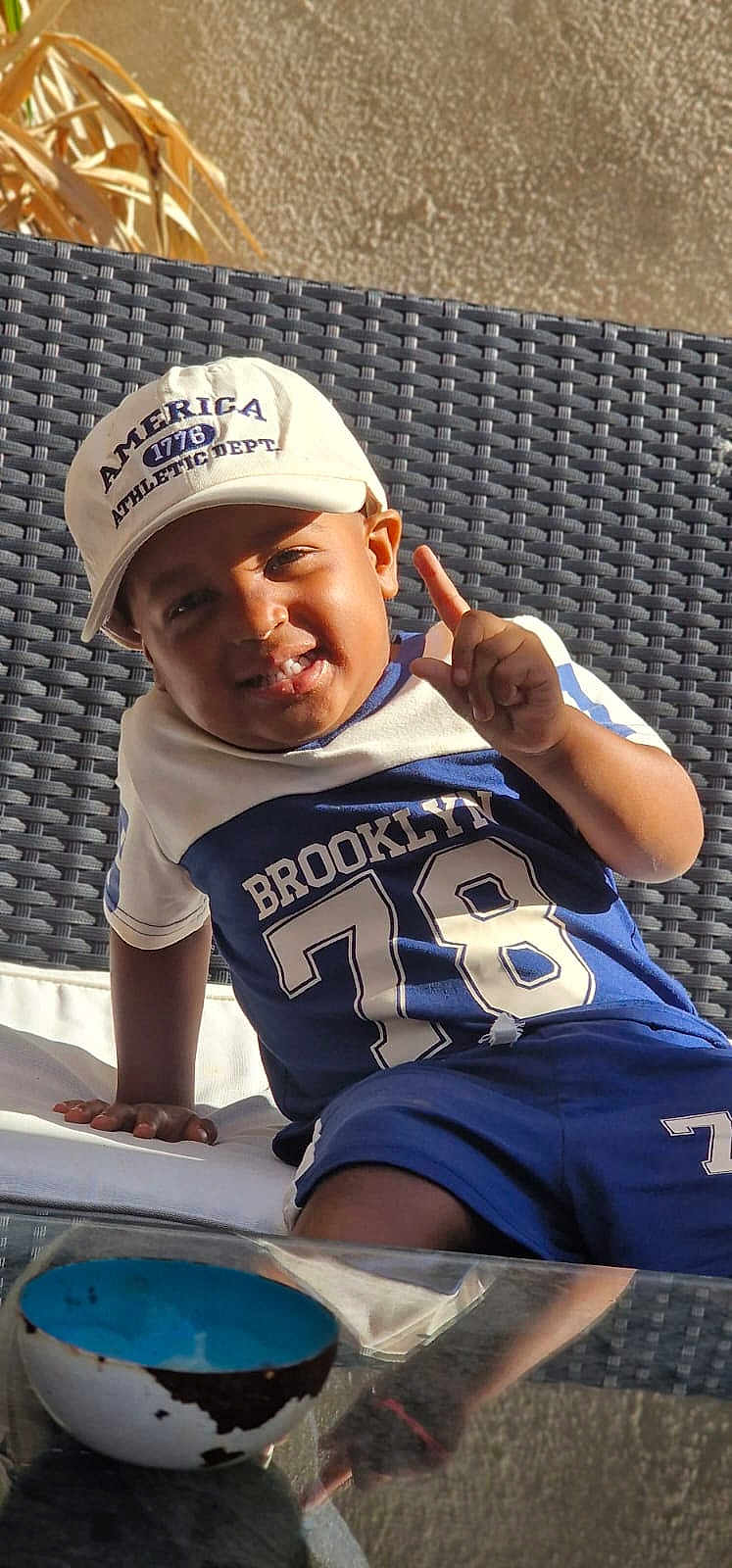 Elvis Johan a rejoint le concours — aidez-le/la à gagner de superbes lots ! blue_clothing, bowl, boy, cap, casual, child, daylight, finger_gesture, glass_table, happy, outdoor, portrait, relaxed, skin, smiling, sportswear, sunlight, toddler, white_cap, woven_chair