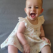 Luana participe au concours pour gagner de l'argent avec cette photo : baby, child, smiling, sitting, dress, white_dress, floral_pattern, happy, indoor, couch, gray_background, cute, infant, person, portrait, barefoot, fun, joyful, young, female