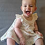 baby, child, smiling, sitting, dress, white_dress, floral_pattern, happy, indoor, couch, gray_background, cute, infant, person, portrait, barefoot, fun, joyful, young, female
