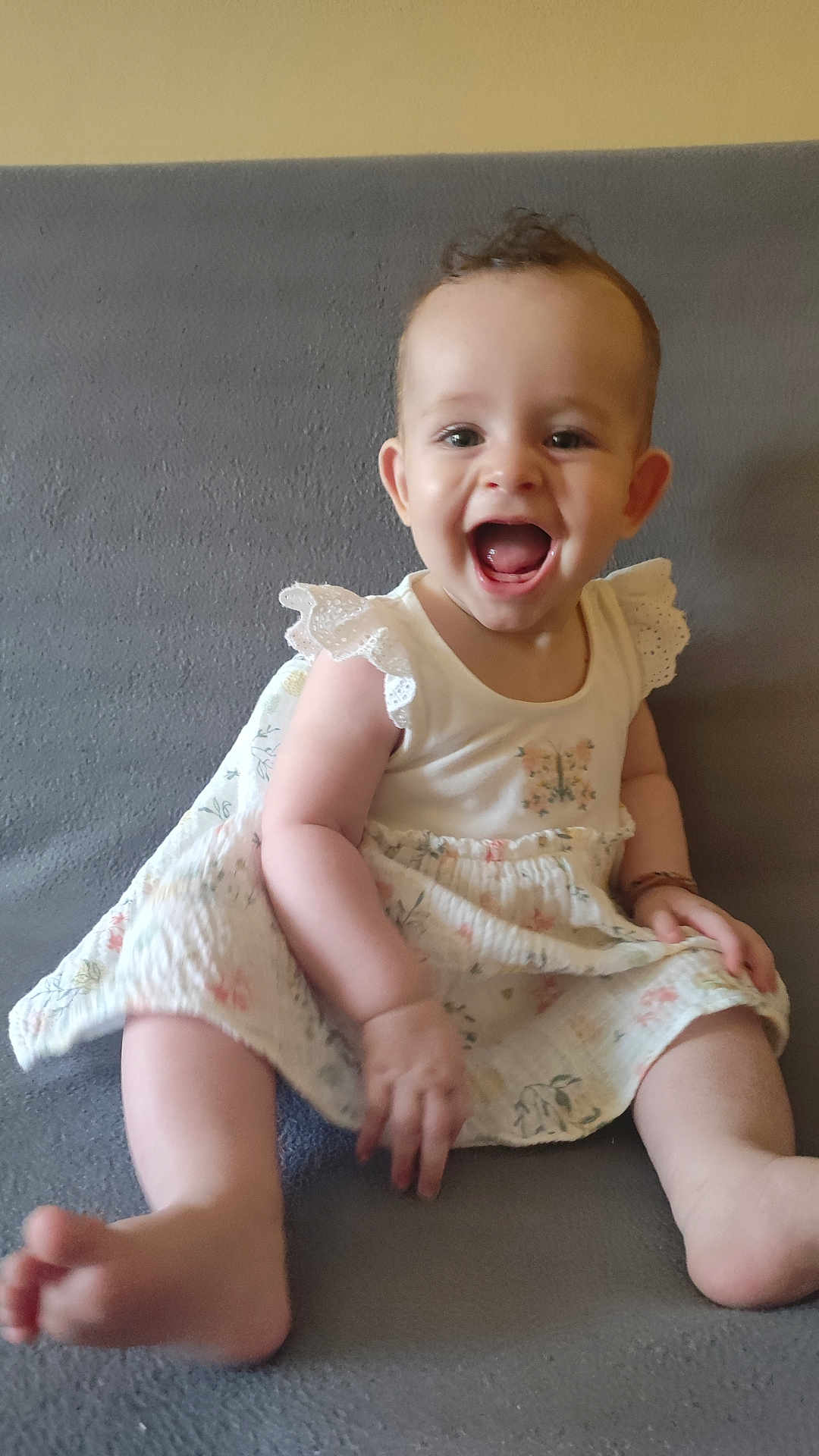 Luana participe au concours pour gagner de l'argent avec cette photo : baby, child, smiling, sitting, dress, white_dress, floral_pattern, happy, indoor, couch, gray_background, cute, infant, person, portrait, barefoot, fun, joyful, young, female