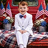 child, boy, white_suit, bow_tie, american_flag, red_truck, plaid_blanket, flowers, patriotic, outdoor, smiling, portrait, sitting, celebration, nature, grass, vintage_vehicle, happy, casual, cute