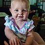 arm, baby, blue_eyes, blurred_background, child, closeup, cute, face, floral_clothing, furniture, hand, happy, holding, indoor, infant, person, sitting, skin, smile, young