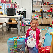 Kadija participe au concours pour gagner de l'argent avec cette photo : child, smiling, unicorn_headband, glasses, pink_vest, tutu, stuffed_unicorn_bag, colorful_case, toys, playroom, gift_wrapping, table, chairs, shelves, clock, dollhouse, handbag, bracelet, floor, happy