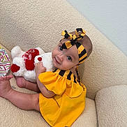 Alana is registered to the contest to win money with this photo: baby, child, plush_toy, yellow_dress, headband, couch, indoor, smiling, cute, infant, soft_toy, red, white, striped, looking_up, seated, happy, cozy, portrait, human