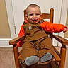 child, toddler, smiling, rocking_chair, wooden_chair, overalls, orange_shirt, carpet, indoor, happy, person, young_child, furniture, portrait, cute, baby, seated, clothing, face, expression