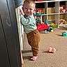 toddler, child, indoor, carpet, toy, playroom, smiling, barefoot, brown_pants, green_sweater, crib, furniture, plastic_toy, colorful, floor_toys, person, young_child, crawling_toy, baby_toy, cute