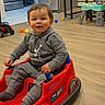 toddler, child, toy_car, red, grey_hoodie, indoor, wooden_floor, toys, balls, smiling, happy, playtime, living_room, furniture, bench, refrigerator, kitchen, home, cute, seat