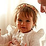 baby, child, infant, smile, face, headband, flower, white_clothing, ruffle, hand, adult_hand, portrait, closeup, warm_light, indoors, cute, happy, toddler, soft_light, gentle