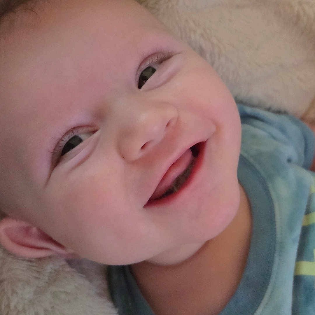 Walker is registered to the contest to win money with this photo: baby, smiling, face, child, infant, blue_shirt, blanket, soft_texture, happy, cute, portrait, closeup, skin, head, eyes, mouth, person, lying_down, cozy, joyful